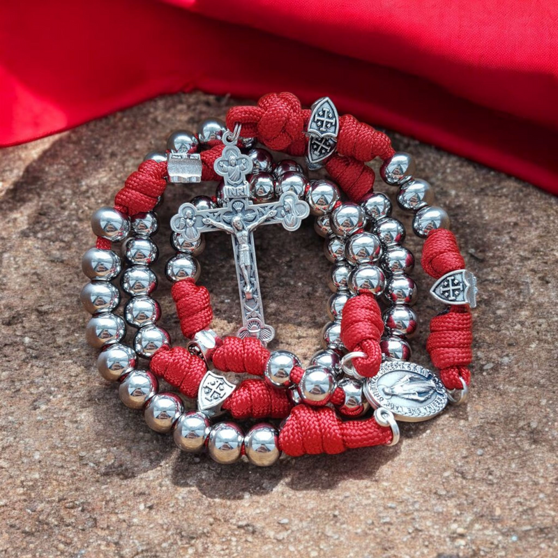 Rosenkranz aus rotem Paracord mit silbernen Edelstahlperlen, Schildperlen mit Jerusalemkreuz und Kruzifix auf sandigem Grund